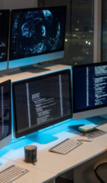 Group of computer monitors with coded data standing in row on desks in empty office of programming company at night time